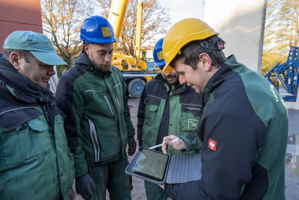 Drei Mitarbeiter in Arbeitskleidung besprechen die Bauplanung am Tablet